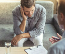 Handsome depressed man in casual clothes is leaning on his knees and telling about his problems while sitting on the couch at the psychotherapist